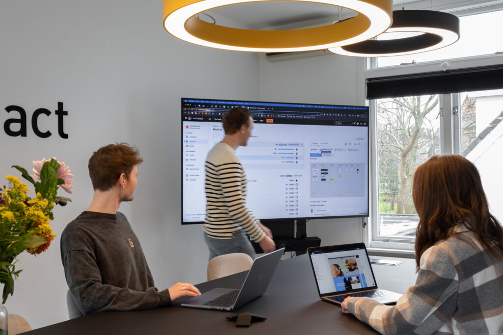 Drie personen zitten rond een tafel in een vergaderruimte. Twee personen kijken naar hun laptops, terwijl de derde persoon voor een groot scherm staat en iets aanwijst. Op het scherm is een interface zichtbaar met projectmanagement software. Er zijn bloemen op tafel en boven hen hangen moderne ronde lampen. De ruimte heeft een grote raam met uitzicht op bomen.